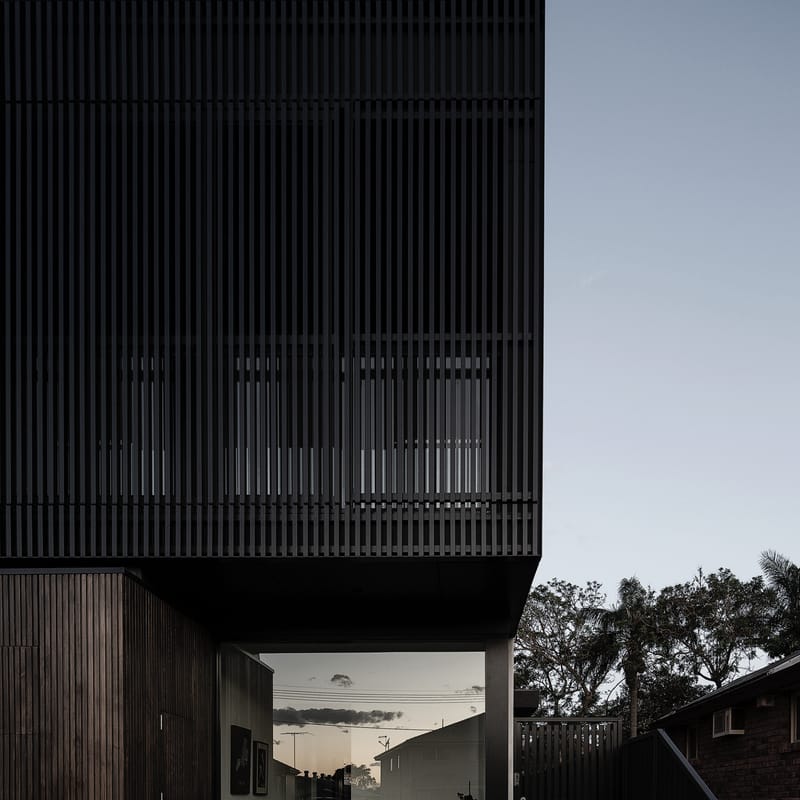 Upper level of a duplex wrapped in black vertical battens over a recessed timber-clad entry, photographed at dusk against a clear sky